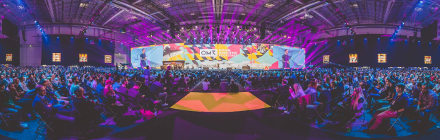 Eine große Menschenmenge, die auf Stühlen sitzt und einer hell erleuchteten Bühne mit mehreren stehenden Individuen zugewandt ist, wahrscheinlich bei einem Online-Marketing-Rockstars-Event.