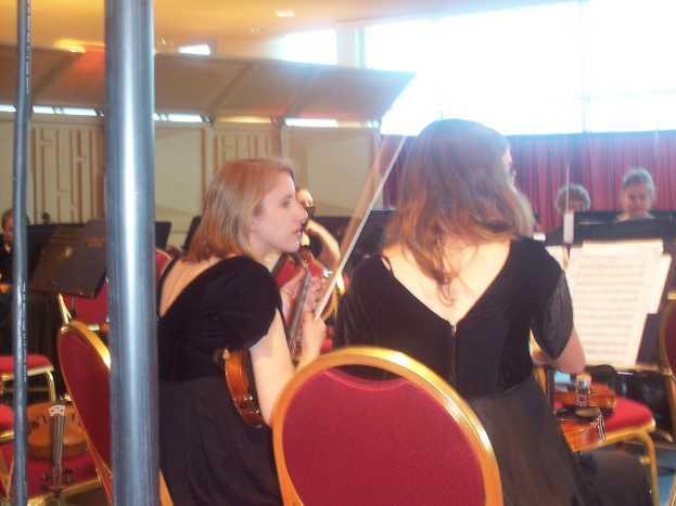 Eine Gruppe von Menschen, die auf Stühlen sitzen, einige halten Musikinstrumente, mit Notenständern, die Notenblätter zeigen, Pfosten und ein Vorhang im Hintergrund.