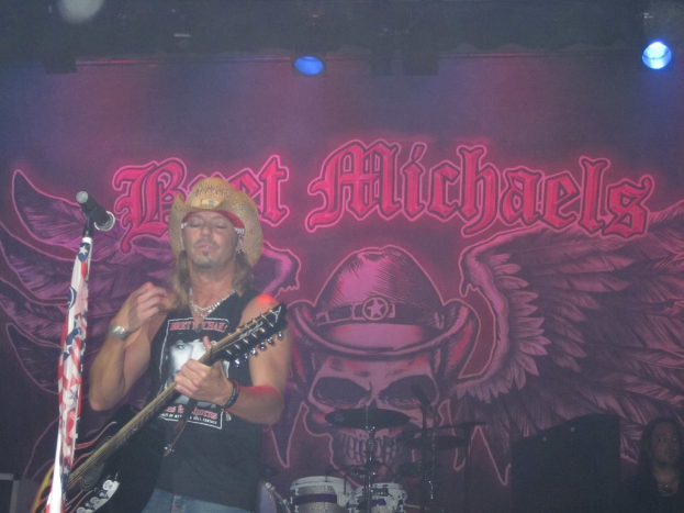 Eine Musik-Konzert-Szene mit einem Gitarristen links neben einem Mikrofon, einer Person in der unteren rechten Ecke, einem Banner im Hintergrund und Drogen in der Mitte unten, alles unter Bühnenbeleuchtung von oben.