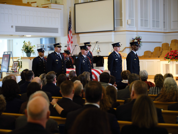 Eine Gruppe uniformierter Personen mit Mützen, die vor sitzenden Trauergästen an einer Beerdigung stehen, mit Blumenbouquets, einer Fahne, einem Bildschirm und einer Wand im Hintergrund.