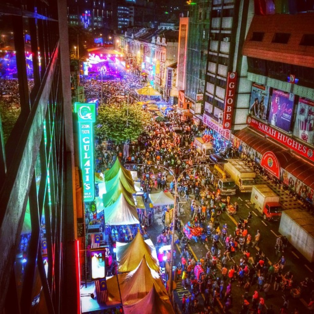 Ein buntes Karnevalsszenario mit einer großen Menschenmenge auf der Straße, umgeben von Zelten, Geschäften, Gebäuden und Bannern, mit zahlreichen Lichtern, die den Hintergrundbereich erhellen.