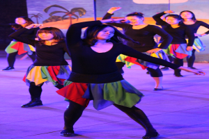 Gruppe junger Frauen in bunten Röcken und Schuhen, die energisch auf einer Bühne tanzen, mit einem Gemälde im Hintergrund.