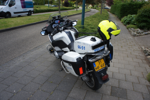 Ein Polizeimotorrad steht am Straßenrand mit einem Helm darauf, umgeben von Bäumen, Pfählen, Gebäuden, Pflanzen, Gras, Steinen und anderen Fahrzeugen im Hintergrund.