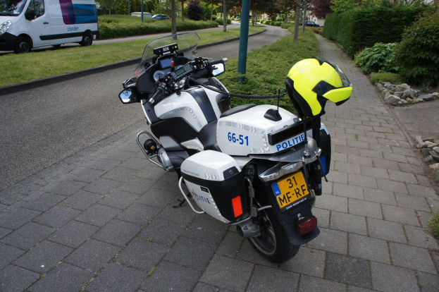 Ein Polizeimotorrad steht am Straßenrand mit einem Helm darauf, umgeben von Bäumen, Pfählen, Gebäuden, Pflanzen, Gras, Steinen und anderen Fahrzeugen im Hintergrund.