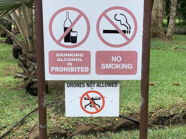 Schilder am Straßenrand mit der Aufschrift "Alkoholkonsum ist untersagt" und "Rauchen verboten" mit Bäumen und Gras im Hintergrund.