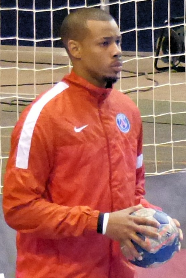 Ein Mann in einem blauen T-Shirt und Shorts steht vor einem Fußballtor und hält einen Fußball mit entschlossener Miene, seine kurzen dunklen Haare und konzentrierten Augen auf den Ball gerichtet.