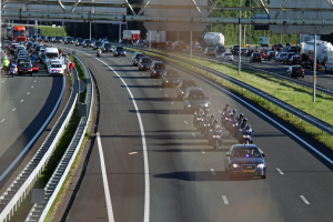 Eine vielbefahrene Autobahn mit zahlreichen Autos und Motorrädern, gesäumt von Geländern und Gras, mit Bäumen, Polen, Texttafeln und einer Brücke mit Stangen im Hintergrund.