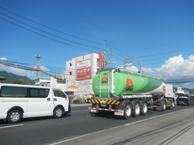 Gro├če Tanklastwagen auf einer Stra├če neben einem wei├čen Lieferwagen mit Strommasten, Geb├Ąuden, Werbetafeln, B├Ąumen, H├Ąngen und einem bew├Âlktem Himmel im Hintergrund.
