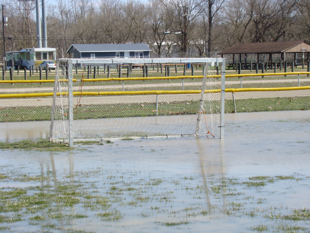 Fußballtor in einem überfluteten Feld mit umliegendem Gras und Wasser, Schuppen, Pfosten, Bäumen, Fahrzeugen und einem klaren blauen Himmel im Hintergrund.