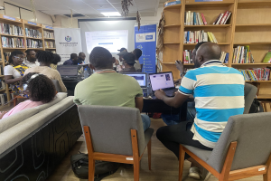 Gruppe von Menschen an einem Tisch mit Laptops in einer Bibliothek, umgeben von Bücherregalen, mit einem Banner, Bildschirm, Deckenbeleuchtung und Taschen auf dem Boden, die auf eine Technologiewerkstatt hinweisen.