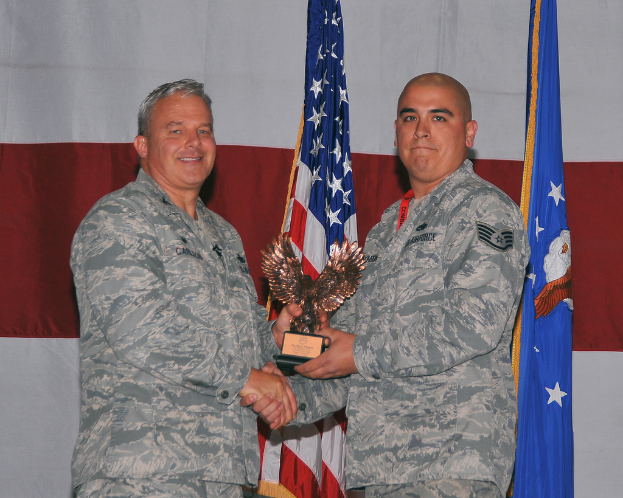 Zwei Personen mit einem Preis stehend, mit Flaggen und einem Vorhang im Hintergrund.