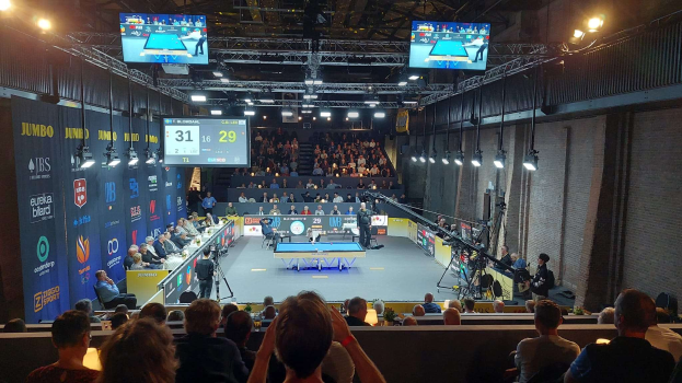 Eine große Menschenmenge sitzt vor einer Bühne mit einem Tischtennistisch in der Mitte, Stühlen auf der linken Seite, einer Wand auf der rechten Seite und Deckenlampen und Fernsehern im Hintergrund bei der 2019 Jumbo-Weltmeisterschaft.