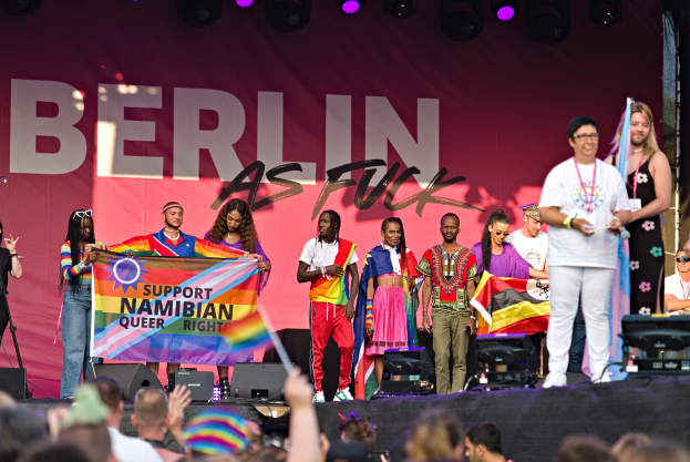 Gruppe von Menschen auf einer Bühne mit einem Banner, auf dem 'Berlin als Fuck' steht, mit Lautsprechern, Mikrofonen und einer Menge unten, die Hände hebt.