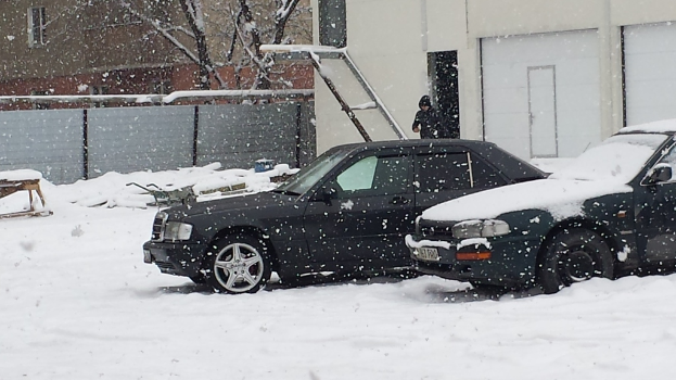 Zwei schneebedeckte Autos in der Nähe von zwei Gebäuden geparkt, mit einer Person daneben und einem Baum zur Seite, alles vor einem schneebedeckten Boden.