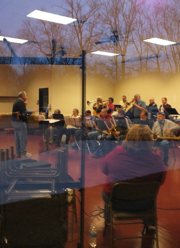 Musiker mit Trompeten im Hintergrund, Sänger vor Mikrofonen und eine stehende Person, die Musik komponiert, mit einer Wand und Bäumen im Hintergrund durch eine Glasreflexion.