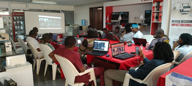 Eine Gruppe von Menschen sitzt um einen Tisch mit Laptops, Mäusen und Flaschen und nimmt an einem digitalen Marketing-Workshop teil, mit einem Banner, einem Display-Bildschirm und elektronischen Geräten im Hintergrund.