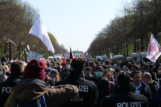 Eine große Gruppe von Menschen steht vor einer Menge von Polizeibeamten, einige tragen Mützen und Masken, während sie auf einer Demonstration in Berlin, Deutschland, mit Schildern, Fahnen, Laternenpfählen, Bäumen, Fahrzeugen, einem Gebäude und dem Himmel im Hintergrund demonstrieren.