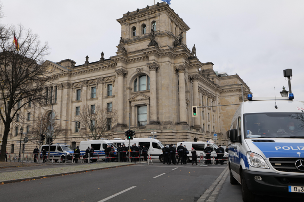 Eine Gruppe von Polizeibeamten steht vor dem Reichstaggebäude in Berlin, Deutschland, mit Fahrzeugen, einem Zaun, Verkehrsampeln, Laternenpfählen, Bäumen und Flaggen im Hintergrund, unter einem klaren Himmel.