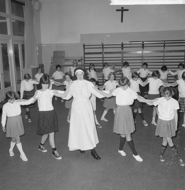 Schwarzes und weißes Bild von Kindern in Schuluniformen, die in einem Raum mit einem Kreuzsymbol an der Wand, Glastüren mit Vorhängen und Deckenlampen tanzen.