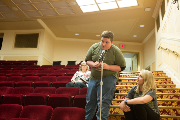 Ein Mann in einem grünen T-Shirt und blauen Jeans steht in einem Theater und hält ein Mikrofon in der Hand, während zwei Frauen auf der Treppe hinter ihm sitzen, rote Stühle im Hintergrund, ein Geländer auf der rechten Seite und Lampen auf dem Dach.