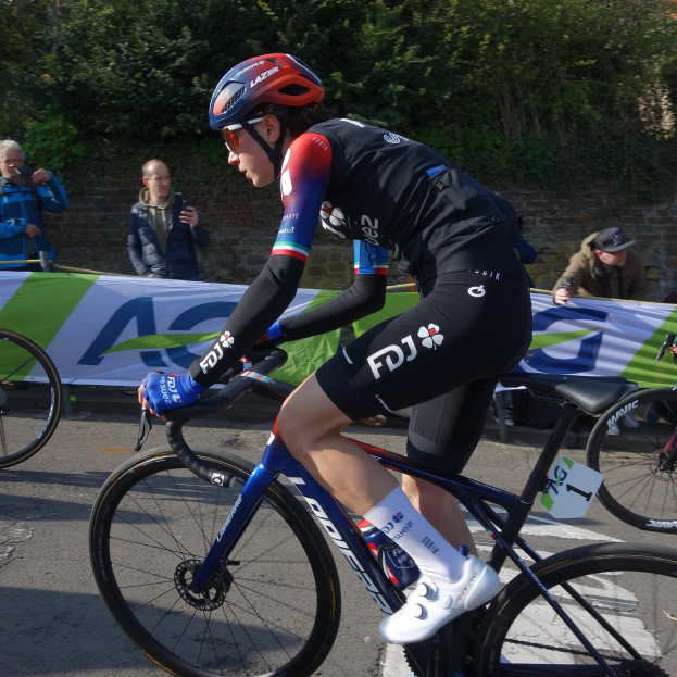 Die professionelle Radrennfahrerin Katie Archibald fährt auf einem Fahrrad die Straße entlang, trägt einen Helm, eine Schutzbrille, Handschuhe und Schuhe und ist von einer Menschenmenge umgeben. Im Hintergrund sind ein Banner, eine Wand, Bäume und ein Gebäude zu sehen.