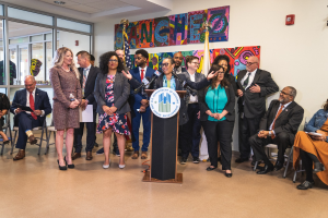 Gruppe von Menschen vor einem Rednerpult mit Mikrofon und einer Tafel mit dem Text "Sancho-Stadtrat tritt dem Stadtrat bei", mit sitzenden und stehenden Teilnehmern, Fahnen, Feuerlöscher und Deckenleuchten im Hintergrund.
