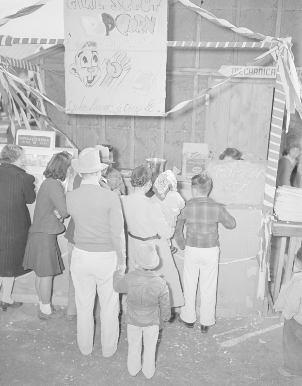 Schwarzes und weißes Foto einer Gruppe von Menschen, die vor einer Karnevalsbude mit Bändern und einer Schautafel stehen.