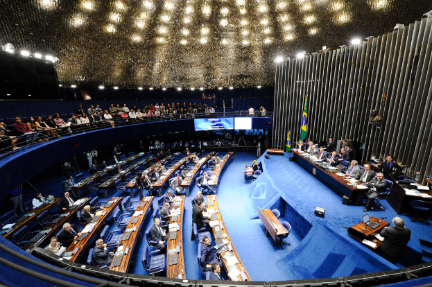 Eine Konferenzbühne mit sitzenden Personen, einem Tisch mit Papieren und Gegenständen, Flaggen und einer Skulptur im Hintergrund, mit Sitzplätzen vor der Bühne und einer Absperrung, beleuchtet von Deckenlampen.