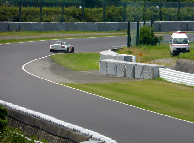 Ein Rennwagen fährt eine Rennstrecke entlang mit einem Polizeiwagen im Hintergrund, umgeben von Gras, Pflanzen, Bäumen und Absperrungen, mit einem Fahrzeug auf der rechten Seite und Text unten.