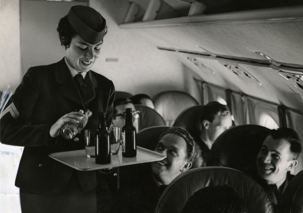 Schwarzes und weißes Bild einer Flugbegleiterin, die Drinks von einem Tablett an sitzende Passagiere auf einem Flugzeug serviert.