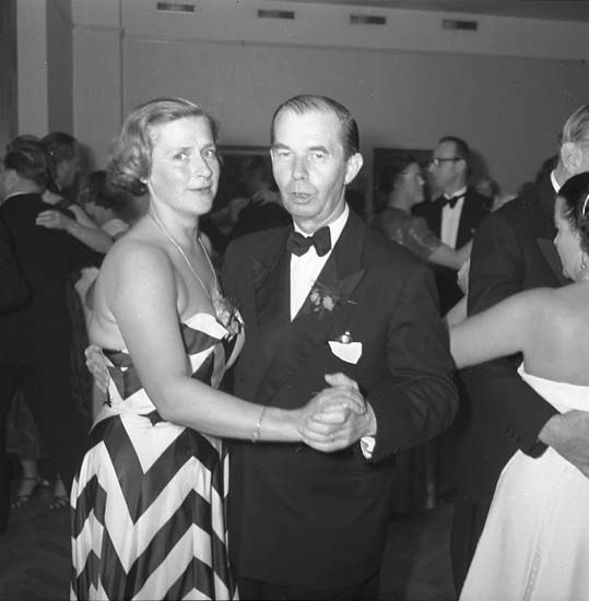 Zwei Tänzer umarmen sich auf einer hell erleuchteten Bühne mit farbenfroher Umgebung, der Mann in einem weißen T-Shirt und blauen Hosen, die Frau in weißen Hosen und einer grünen Oberteil.