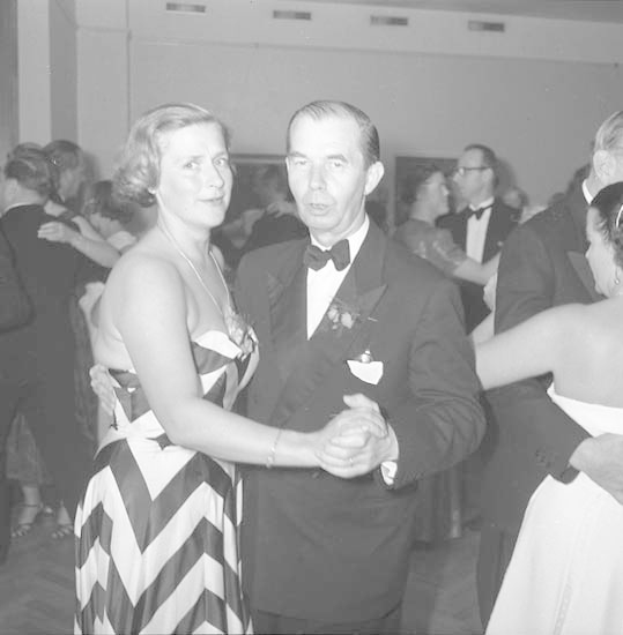 Zwei Tänzer umarmen sich auf einer hell erleuchteten Bühne mit farbenfroher Umgebung, der Mann in einem weißen T-Shirt und blauen Hosen, die Frau in weißen Hosen und einer grünen Oberteil.