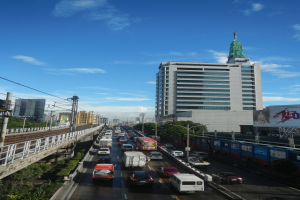 Eine belebte Stadtstraße mit starkem Verkehr, hohen Gebäuden, einer Brücke auf der linken Seite, Strommasten mit Kabeln, Bäumen und einem bewölkten Himmel im Hintergrund.