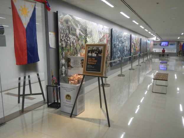 Langer Flur mit einer philippinischen Flagge an der Wand, Displaytafeln mit Text und Bildern, Ribbonständer, Bänke, Deckenleuchten und ein Bildschirm im Hintergrund, der wahrscheinlich Informationen über das Philippinische Nationalmuseum anzeigt.