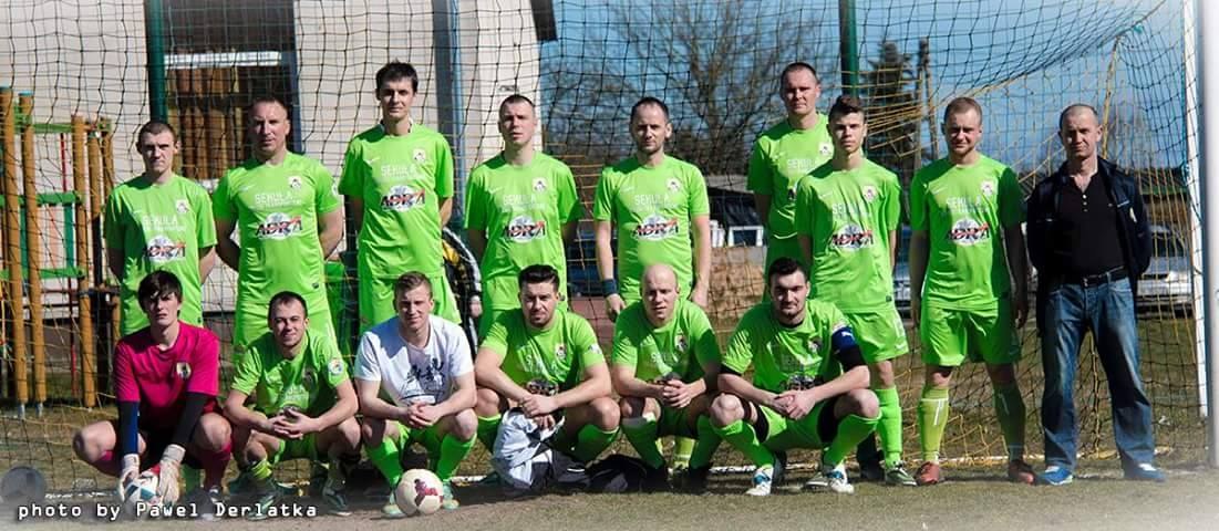 Eine Gruppe von Männern in grünen T-Shirts steht auf einem Fußballfeld neben einem Torpfosten, mit einem Ball auf dem Boden und verschiedenen Hintergrundelementen wie Bäume, Gebäude und Fahrzeuge.