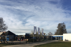Ein große Gebäude mit einem grünen Bereich davor, umgeben von einem Metallzaun, einem Weg, Bäumen, Pfosten, Bannern, einem geparkten Fahrrad, nächsten Häusern und einem bewölktem Himmel, das als neuer Spielplatz an einer Schule dient.