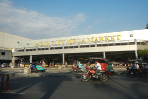 Ein belebter Pasig City Mega Market mit Fahrzeugen auf der Straße, Menschen, die gehen, einem Gebäude mit Text im Hintergrund und einem bewölkten Himmel.