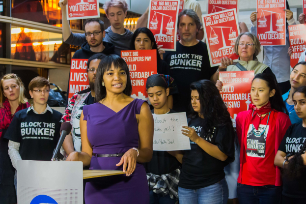 Eine Black-Lives-Matter-Aktivistin spricht an einem Podium mit Mikrofon vor einer Menge mit Schildern, mit Lichtern und Gegenständen im Hintergrund.