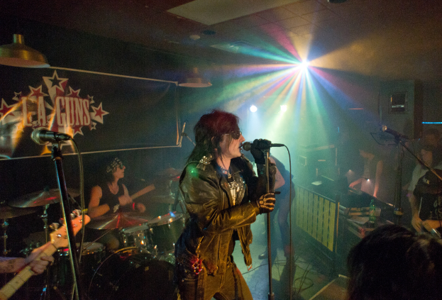 Eine Frau in einer schwarzen Jacke singt in ein Mikrofon auf der Bühne, mit einem Mann in Sonnenbrille und Headband, der hinter ihr ein Instrument spielt, und einem Plakat mit der Aufschrift "LA Guns" im Hintergrund, unter Diskoleuchten und hängenden Lichtern.