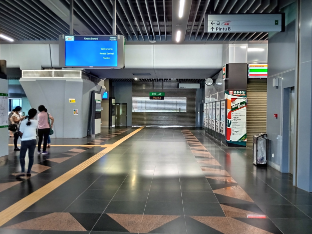 Eine Gruppe von Menschen mit Taschen geht durch einen Bahnhof, mit einem Gepäckwagen auf der rechten Seite, Texttafeln, eine Wanduhr und Deckenbeleuchtung.