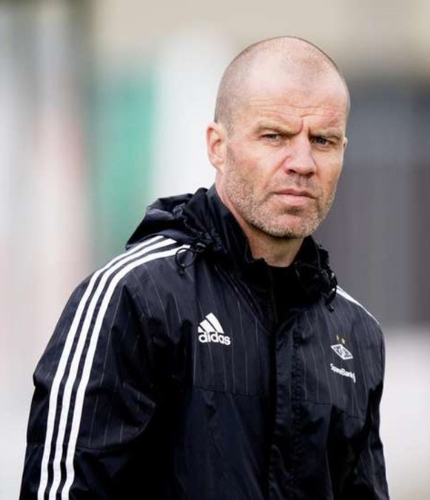 Ein kahlköpfiger Trainer in einer blauen Jacke mit weißen Streifen und einem weißen Kragen steht auf einem Fußballfeld mit verschränkten Händen und schaut ernst nach vorne.