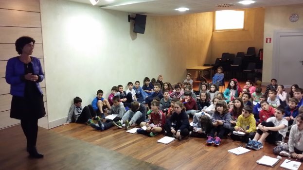 Eine Gruppe von Kindern, Jungen und Mädchen, sitzt auf dem Boden mit Büchern vor sich, während eine Frau auf der linken Seite auf einer Bühne steht, mit einer Wand im Hintergrund.