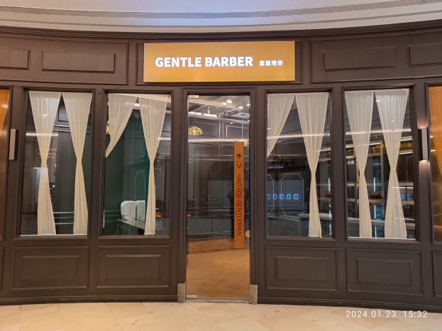 Innenansicht eines Friseursalons in einem Einkaufszentrum mit Glasfenstern, Vorhängen, einer Wandtafel, Deckenleuchten und einem Wasserzeichen.