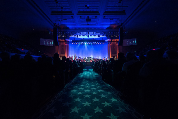 Ein großes Auditorium mit Sitzplätzen, eine Bühne mit Musikern und über Kopfbeleuchtung und -bildschirme, was auf einen Konzertsaal bei der Nationalen Convention 2019 hindeutet.