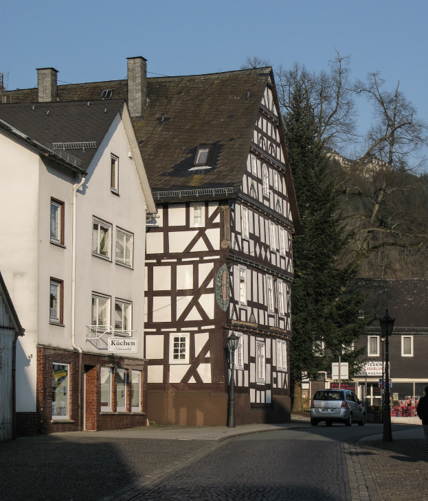 Haus aus Fachwerk in einer kleinen Stadt umgeben von Gebäuden, Fahrzeugen, Fußgängern, Bäumen und Straßeninfrastruktur unter einem klaren blauen Himmel.