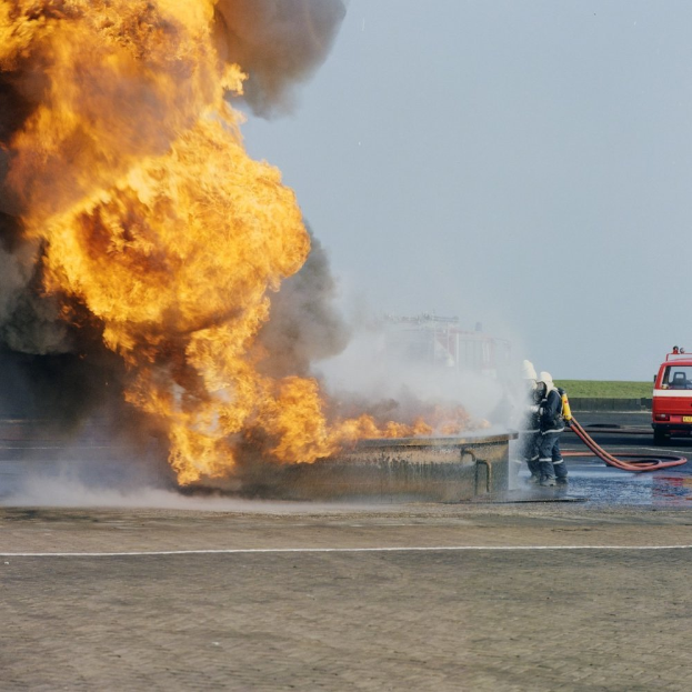 Ein Feuerwehrauto steht in Flammen am Straßenrand, zwei Personen mit Helmen halten Schläuche in der Nähe und ein Fahrzeug und der Himmel sind im Hintergrund zu sehen.
