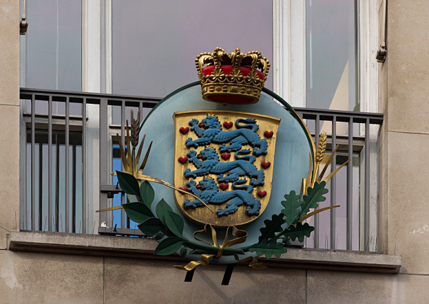 Wappen der britischen Königsfamilie an einer Gebäudewand mit einer Krone- und Blätter-Logo, Fenstern und Metallgittern im Hintergrund.