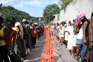 Eine lange Schlange von Menschen, die vor einem Zaun auf ihre Abstimmung warten, mit Bäumen, Gebäuden, Pfählen, Drähten und einer Wand im Hintergrund.