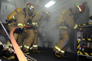 Feuerwehrleute in Schutzausrüstung mit Helmen und Sauerstoffflaschen arbeiten an einem Feuerwehraufsatz, mit einem Gestell voller Gegenstände und Hintergrundrohren und -leuchten.