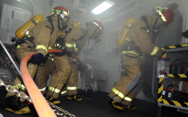 Feuerwehrleute in Schutzausrüstung mit Helmen und Sauerstoffflaschen arbeiten an einem Feuerwehraufsatz, mit einem Gestell voller Gegenstände und Hintergrundrohren und -leuchten.